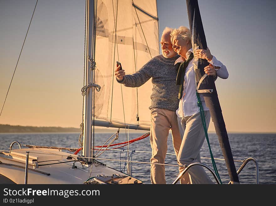 Beautiful senior couple hugging and smiling while making selfie on smartphone, they are standing on the side of sailboat or yacht deck floating in sea, sailing together. Boat trip, travelling by sea