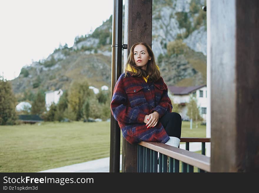 Young woman in coat sitting on wooden fence near cottage on the mountains background