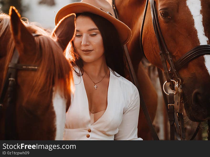 A girl in a white sundress and hat stands next to horses in nature in the eagle stables.