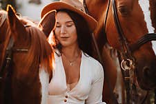 A Girl In A White Sundress And Hat Stands Next To Horses In Nature In The Eagle Stables Royalty Free Stock Image