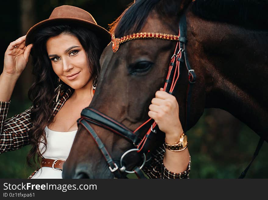 Pregnant girl with a big belly in a hat next to horses in the forest in nature.Stylish girl in white clothes and a brown jacket.