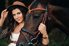 Pregnant Girl With A Big Belly In A Hat Next To Horses In The Forest In Nature.Stylish Girl In White Clothes And A Brown Jacket Royalty Free Stock Photo