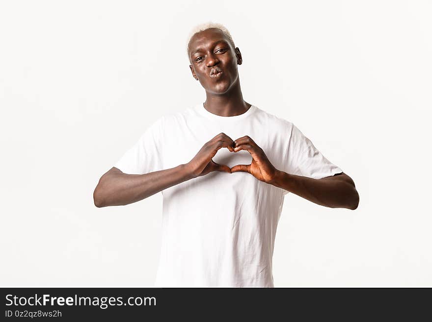 Portrait of cute african-american blond man, pouting for kiss and showing heart gesture, express love, white background