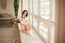 Girl In A White Sweater Dress Against The Background Of A Large Window In A Rustic Style. Young Woman At Home On The Windowsill Fo Stock Photos