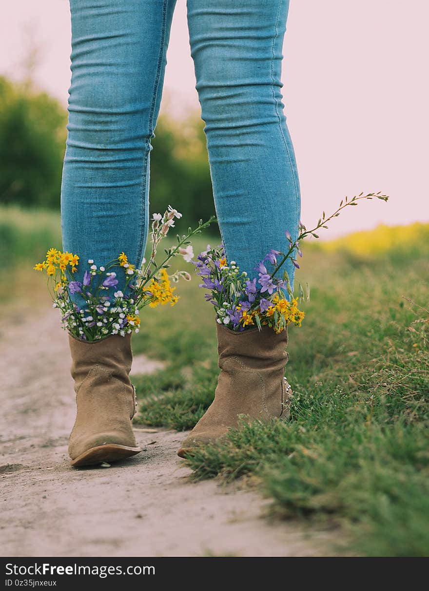 Female legs in jeans high boots with inserted flowers on the road in the field