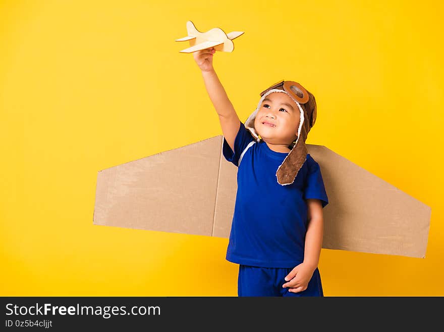 Happy Asian handsome funny child or kid little boy smile wear pilot hat play and goggles with toy cardboard airplane wings fly hold plane toy, studio shot isolated yellow background, Startup freedom