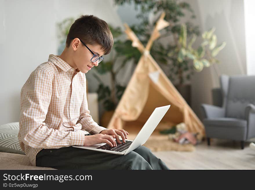 Teenager boy with glasses works in his laptop against the background of his room