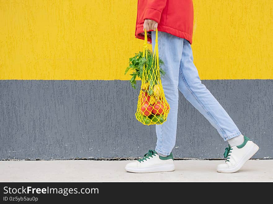 Yellow string bag with cucumbers, tomatoes, bananas and herbs in hands of girl in blue jeans, red jacket. Bright photo in red, yellow and green tones. Sustainability, zero waste, plastic free concept