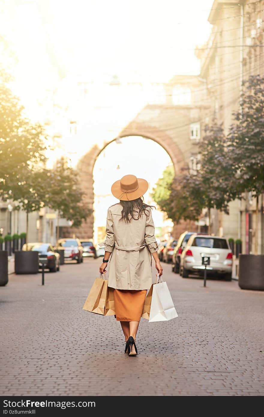 Rear view of a stylish woman wearing grey coat and hat with shopping bags walking city streets, full length