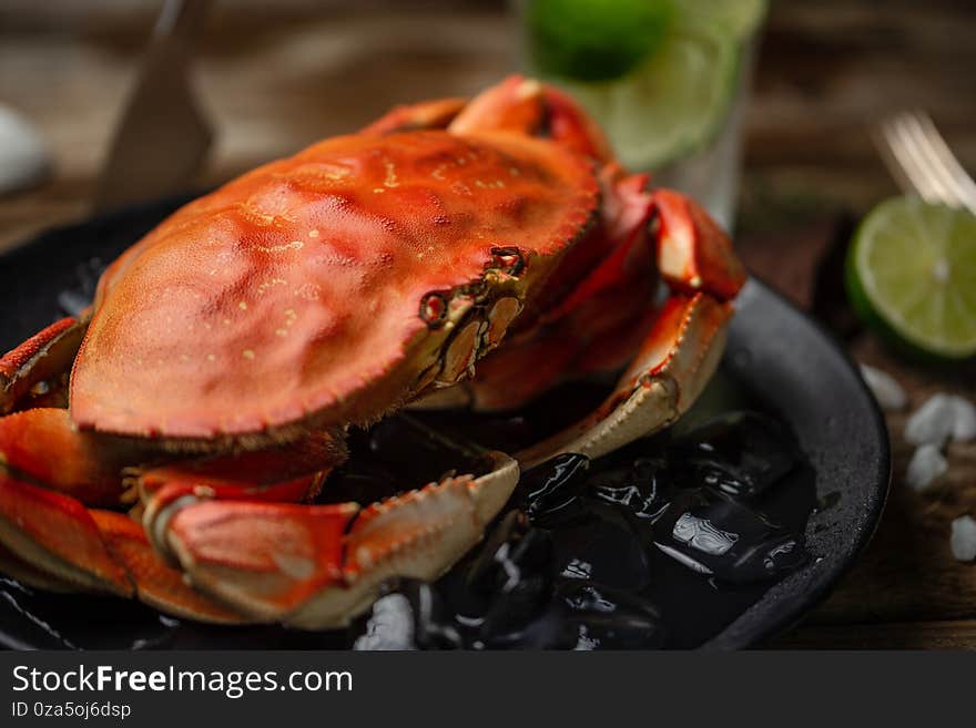 Close-up view of tasty cooked crab on black round plate with ice cubes served with lime and seashells on rustic wooden table, glass of mojito background. Delicious meal. Seafood concept. Horizontal.