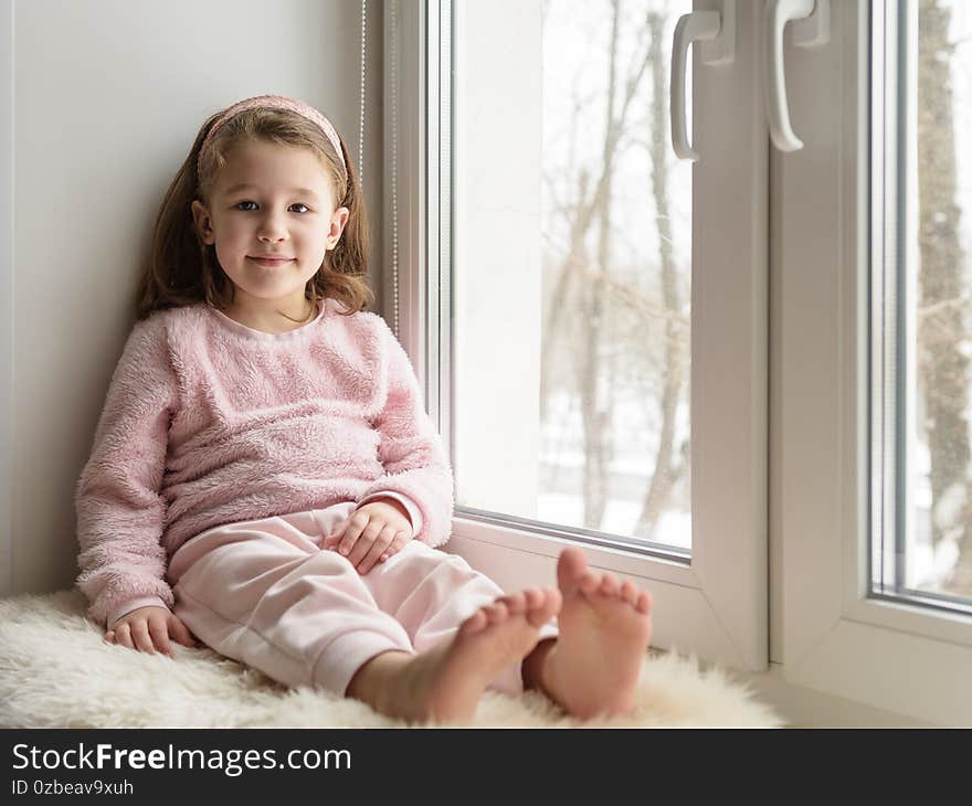 Kid sitting by window looks at camera indoor, portrait of pretty little girl on fur rug on room sill in winter