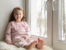 Kid Sitting By Window Looks At Camera Indoor, Portrait Of Pretty Little Girl On Fur Rug On Room Sill In Winter Stock Photography