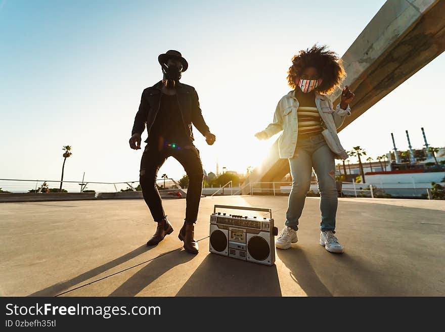 Young Afro friends wearing face mask dancing outdoor while listening to music with wireless headphones and boombox