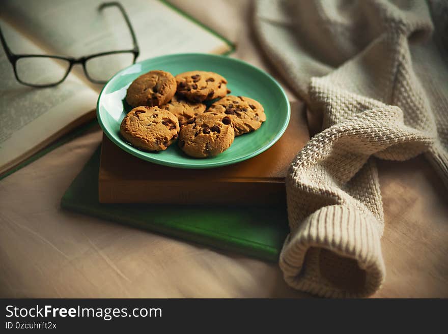 A green plate of chocolate homemade cookies are on top of a stack of old books, next to an open book that has recently been read and a thick warm sweater. Leisure on cold days. The comfort of home. A green plate of chocolate homemade cookies are on top of a stack of old books, next to an open book that has recently been read and a thick warm sweater. Leisure on cold days. The comfort of home