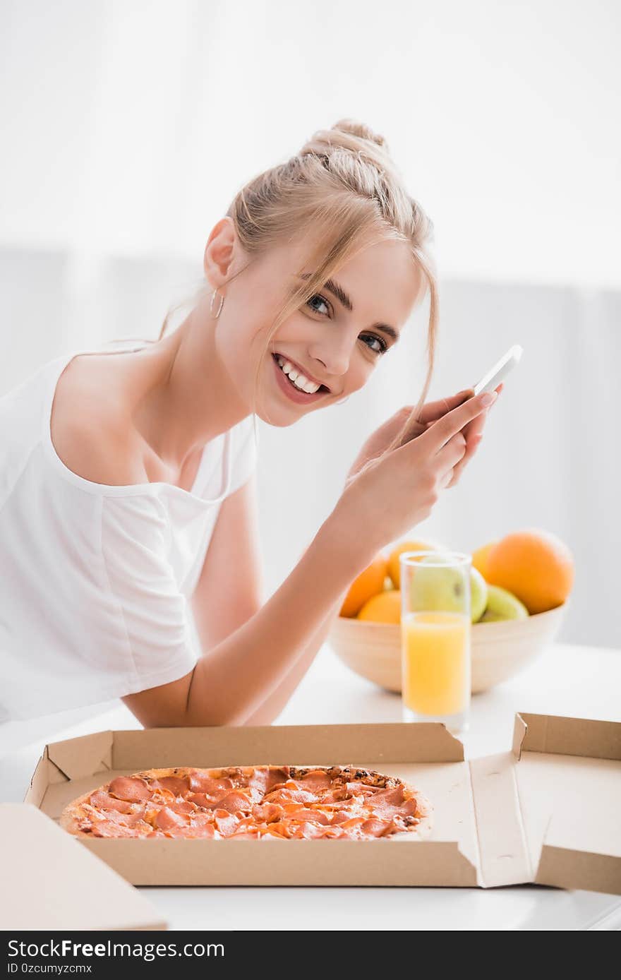 Pleased blonde woman looking at camera while chatting on smartphone in kitchen