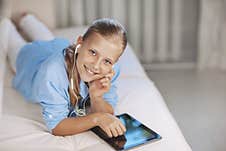 Blonde Teenage Girl Lying On The Couch Plays A Game On The Tablet And Communicates Via Video Link With Friends. Digital Stock Photo