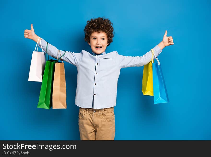 Excited and curly boy standing with outstretched hands and holding shopping bags on blue