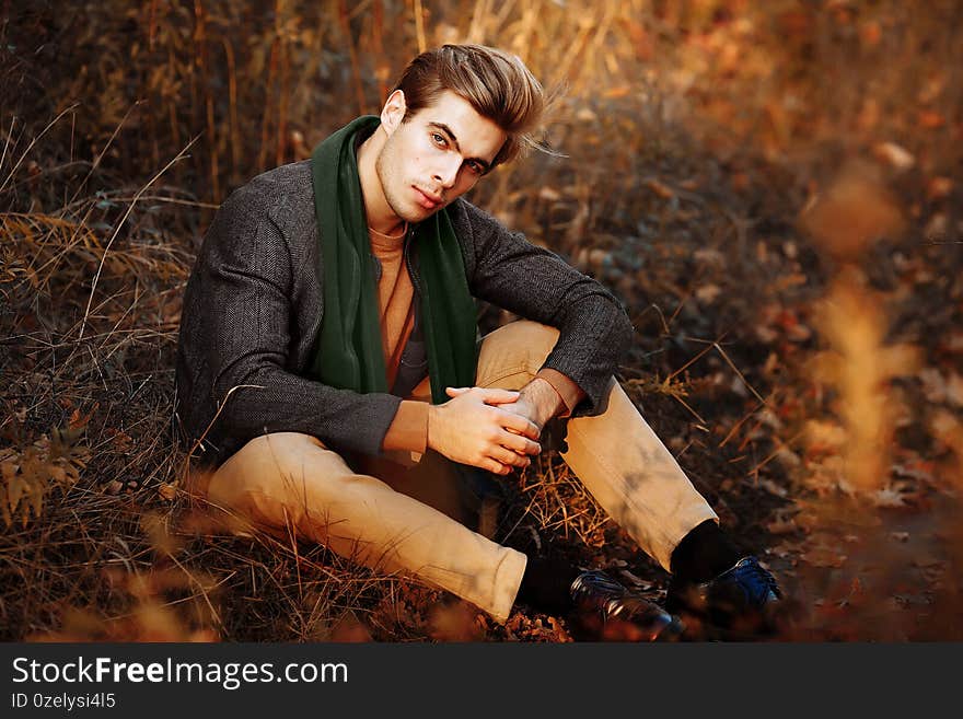 A man, a model in a gray jacket, a green scarf and beige pants and a sweater sits on the grass in autumn, among the autumn trees. posing. thoughtfulness and sadness. male power