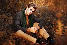 A Man, A Model In A Gray Jacket, A Green Scarf And Beige Pants And A Sweater Sits On The Grass In Autumn, Among The Royalty Free Stock Photo