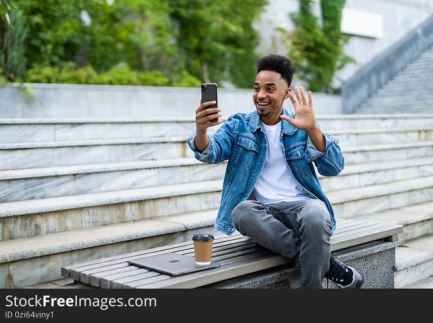 Smiling young african man in casual clothes making video call with smartphone and gesturing greeting while sitting on bench in