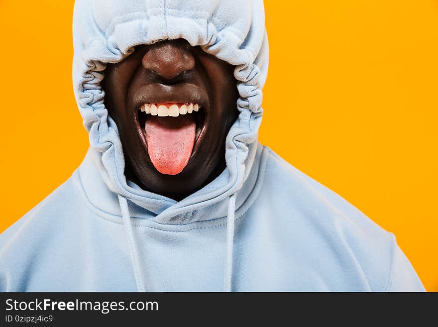 Image of funny african american guy in streetwear hoodie sticking out his tongue isolated over yellow background. Image of funny african american guy in streetwear hoodie sticking out his tongue isolated over yellow background