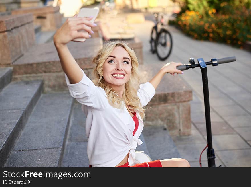 Young blond woman, sitting on staircase of old historical building in city center, taking picture with her phone. Traveler