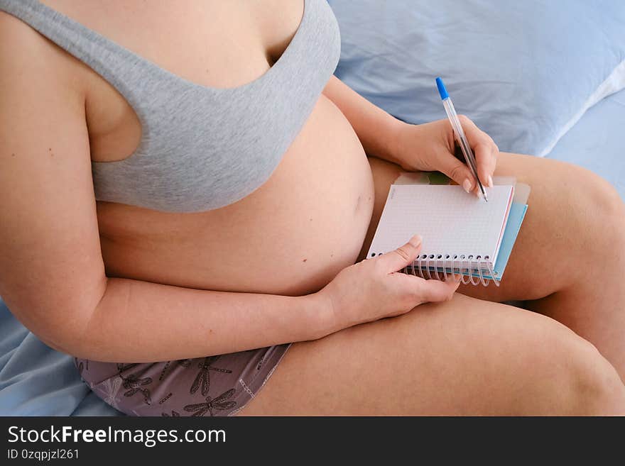 Close-up of a pregnant woman making a packing list for a maternity hospital at home. The expectant mother is engaged in planning, writes shopping lists in a notebook while sitting on the bed. Close-up of a pregnant woman making a packing list for a maternity hospital at home. The expectant mother is engaged in planning, writes shopping lists in a notebook while sitting on the bed