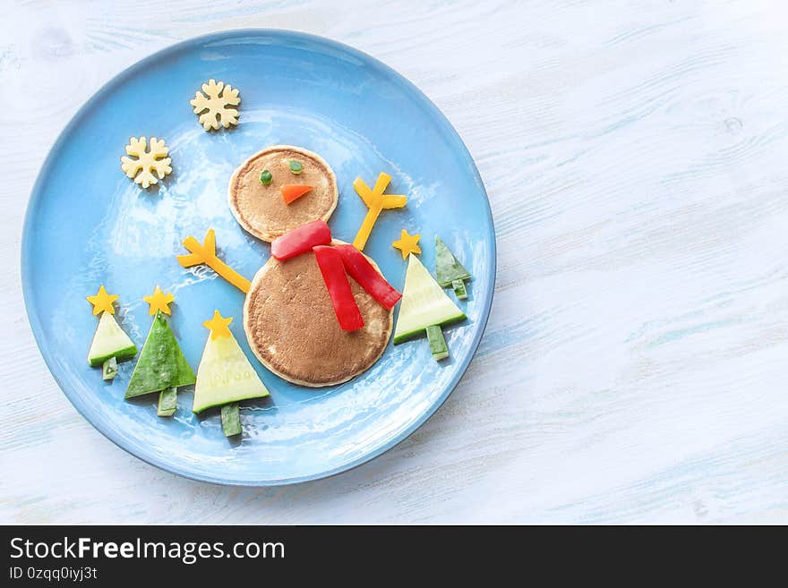 Christmas pancake shaped like a snowman with fresh vegetables blue plate on wooden white table. Christmas fun food for kids, funny breakfast brunch idea xmas. Christmas pancake shaped like a snowman with fresh vegetables blue plate on wooden white table. Christmas fun food for kids, funny breakfast brunch idea xmas