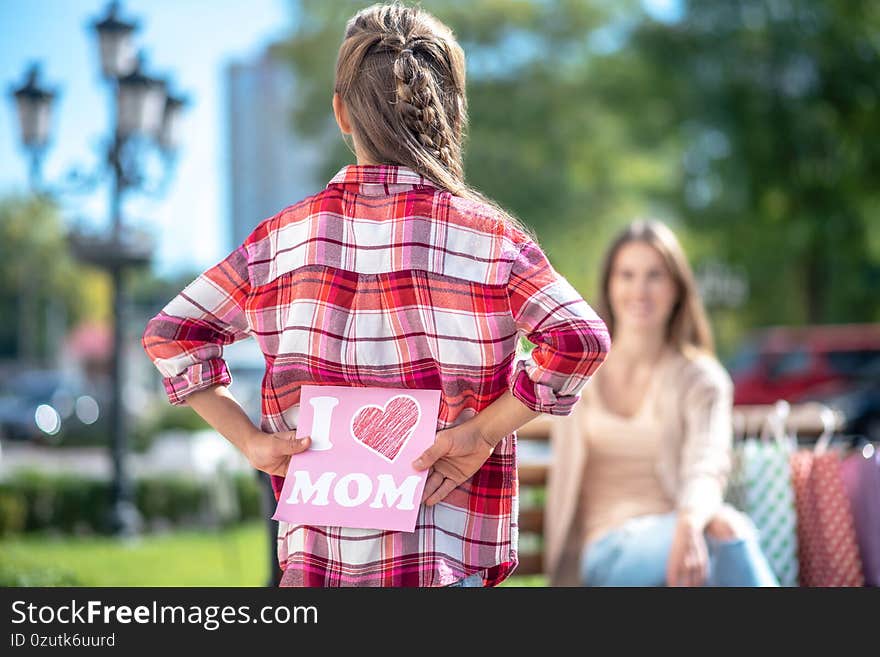 Mom and daughter. Rear view of girl holding card for her mom sitting on park bench