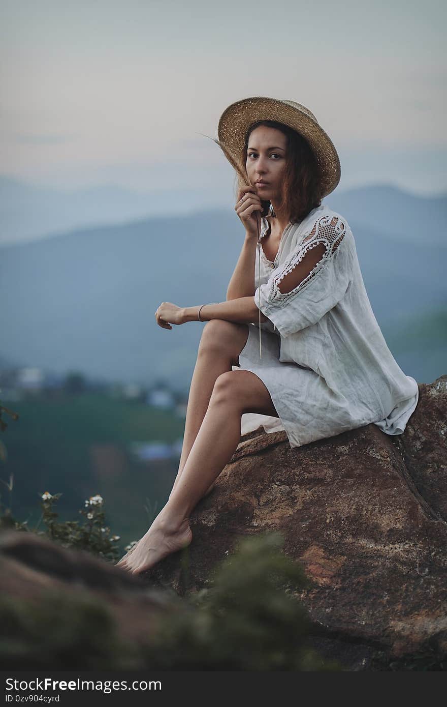 The blonde in a linen dress and straw hat sits on a viewpoint in the mountains