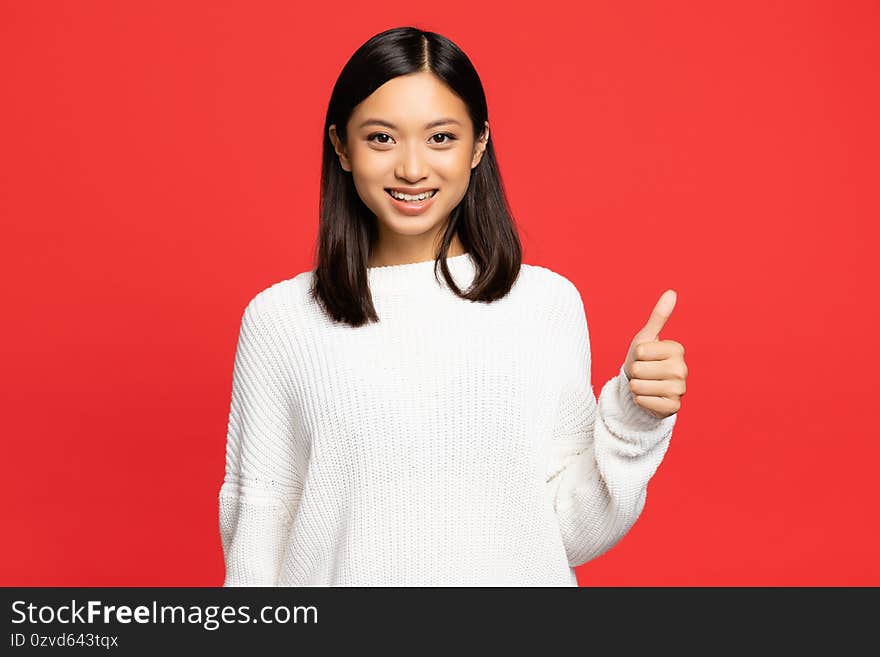 Happy asian woman in sweater showing thumb up isolated on red