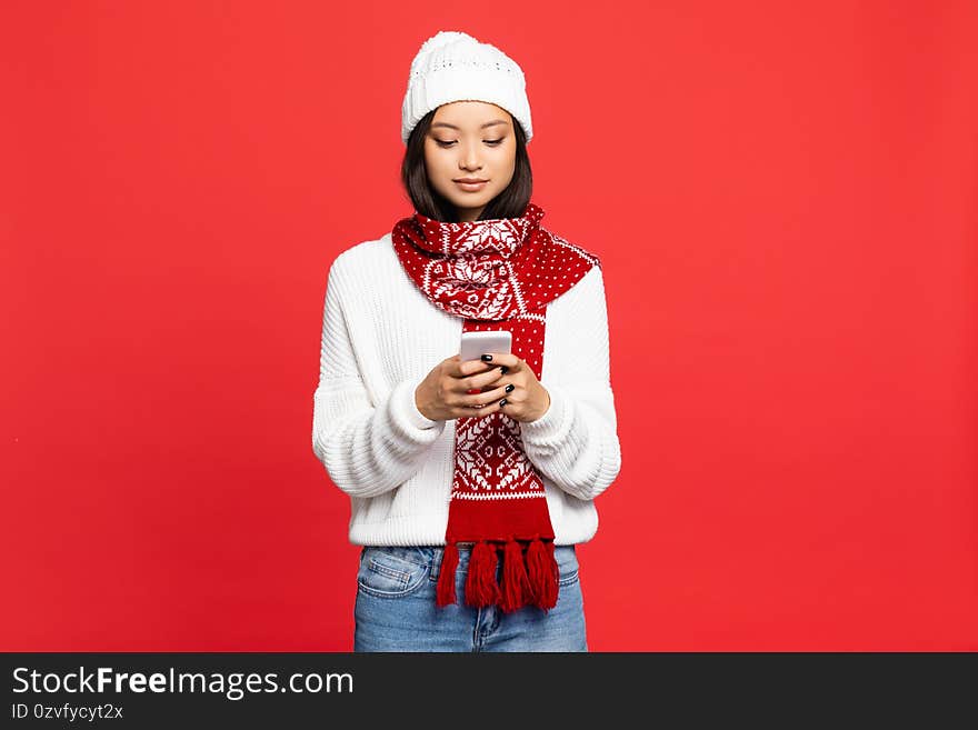 Young asian woman in hat and scarf texting on smartphone isolated on red