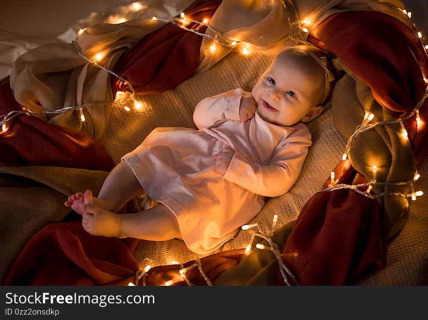 A little three-month-old girl in a pink dress lies in the fabric among the yellow lights and smiles. New Year