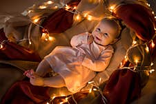 A Little Three-month-old Girl In A Pink Dress Lies In The Fabric Among The Yellow Lights And Smiles. New Year Royalty Free Stock Photography