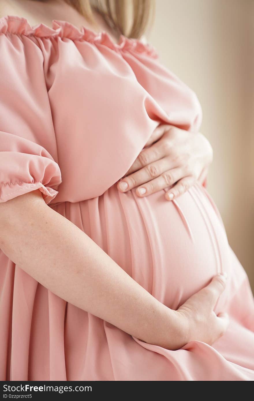 Pregnant woman at home in pink dress hugs her belly