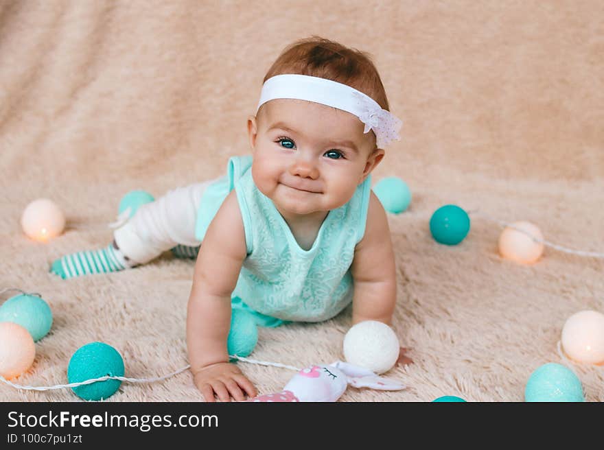 Cute smiling little girl with beautiful blue eyes in blue dress is sitting on a beige plush plaid with Christmas lights before