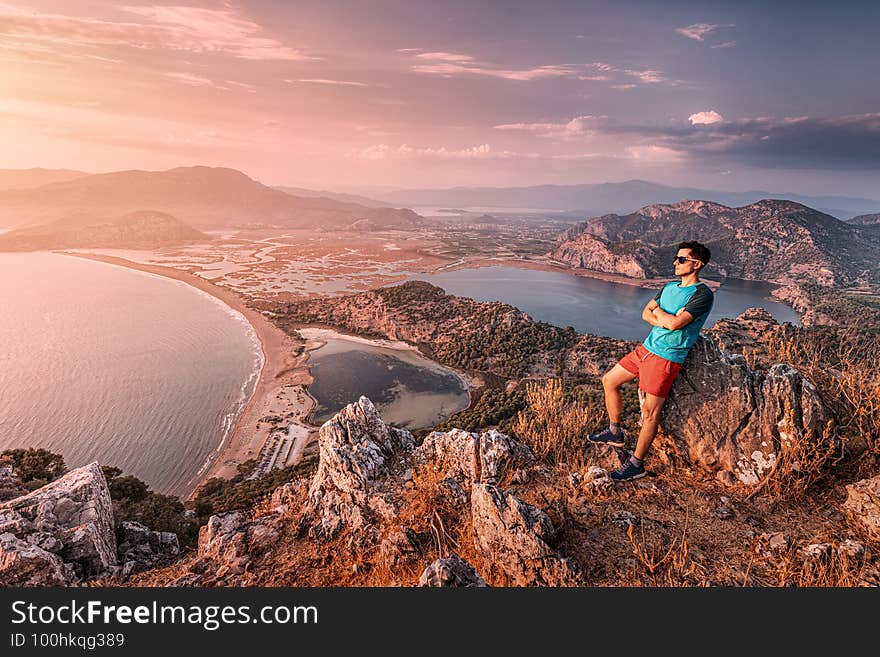 Confident man in sunglasses and red shorts surveys the surrounding area from a viewpoint. In the background, a colorful sunset