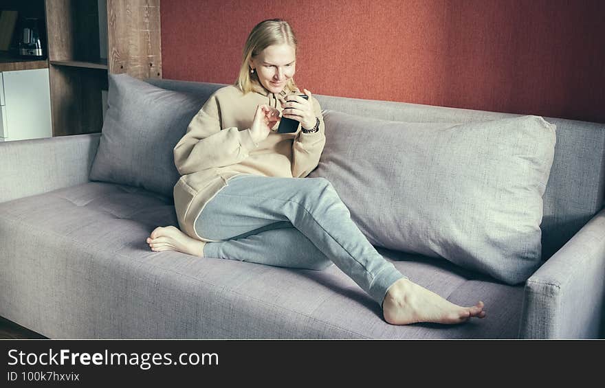 Young woman looking at her smart phone and smiling while sitting on the sofa at home. Woman in the sun