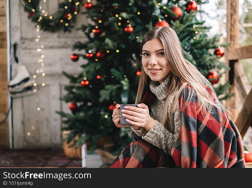 Young caucasian woman in a plaid is drinking a hot drink on the veranda with Christmas tree and decorations