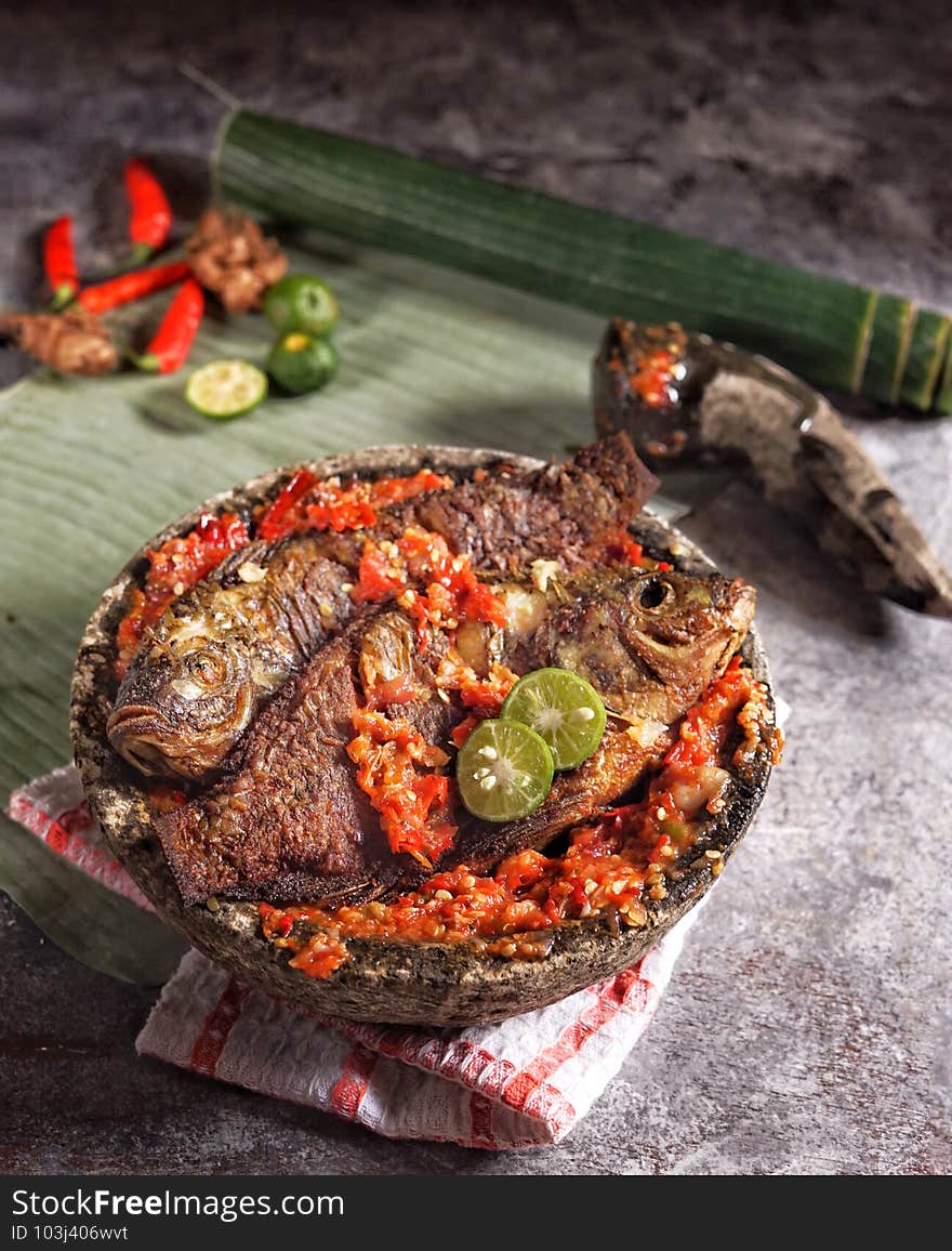 The spicy tilapia is served in a mortar, base of banana leaves. Fried or grilled tilapia fish spiked with lime sauce.  Sundanese, West Java, Indonesian traditional cuisine. The spicy tilapia is served in a mortar, base of banana leaves. Fried or grilled tilapia fish spiked with lime sauce.  Sundanese, West Java, Indonesian traditional cuisine.