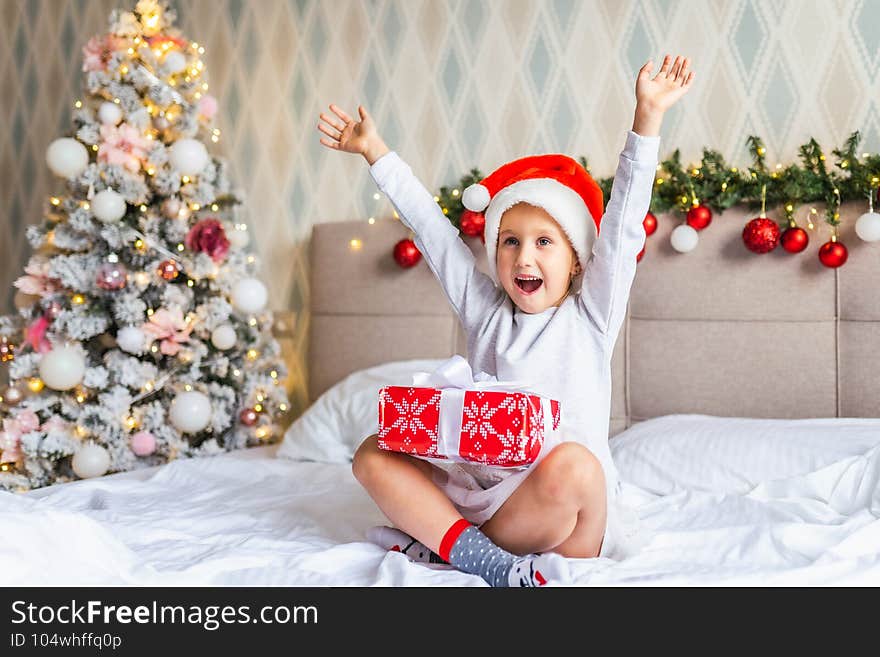 Preschool funny girl sit on bed near decorated Christmas tree in beautiful room in the holiday morning, enjoing with