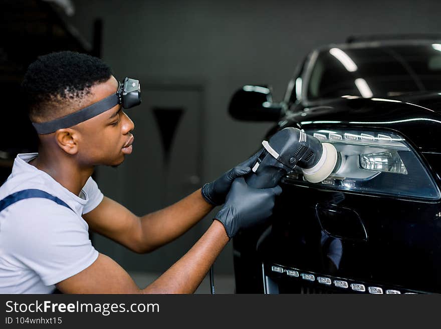 Detailing and polishing of car headlight. Young dark skinned worker in uniform and protective gloves, with orbital polisher in auto service polishing car lights.