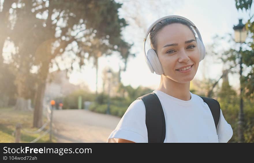 Portrait of beautiful girl in headphones with backpack smiling on camera on the way to morning training in city park