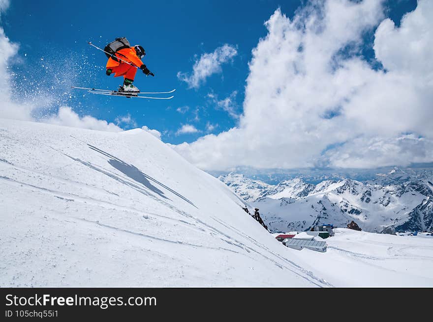 Man skier athlete makes a jump in flight on a snowy slope against the backdrop of a blue sky of mountains and clouds