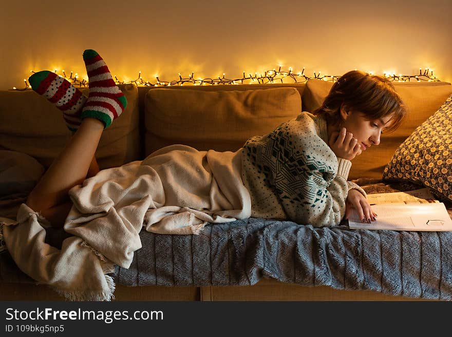 Christmas theme, mood, woman with green and red socks under beige blanket laying on comfy sofa and reading magazine, short blond hair, caucasian, warm tones, christmas lights. Christmas theme, mood, woman with green and red socks under beige blanket laying on comfy sofa and reading magazine, short blond hair, caucasian, warm tones, christmas lights