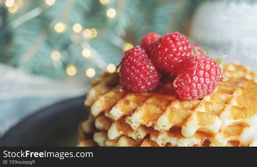 Viennese waffles with raspberries and honey on the Christmas table with tree branches and garlands of lights in the background. close-up, copy space.