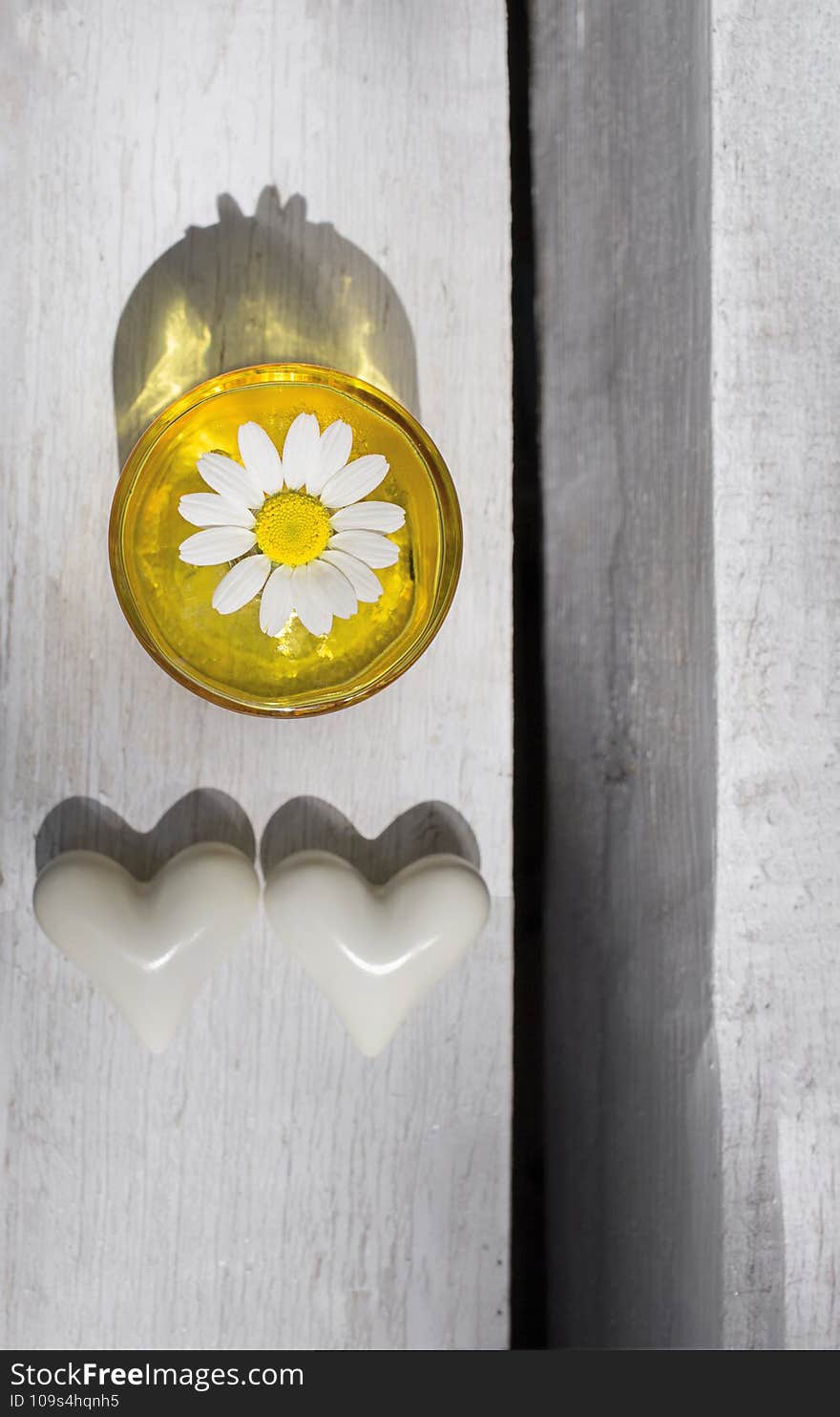 On light wooden background, yellow glass vase with white chamomile flower and two white ceramic hearts