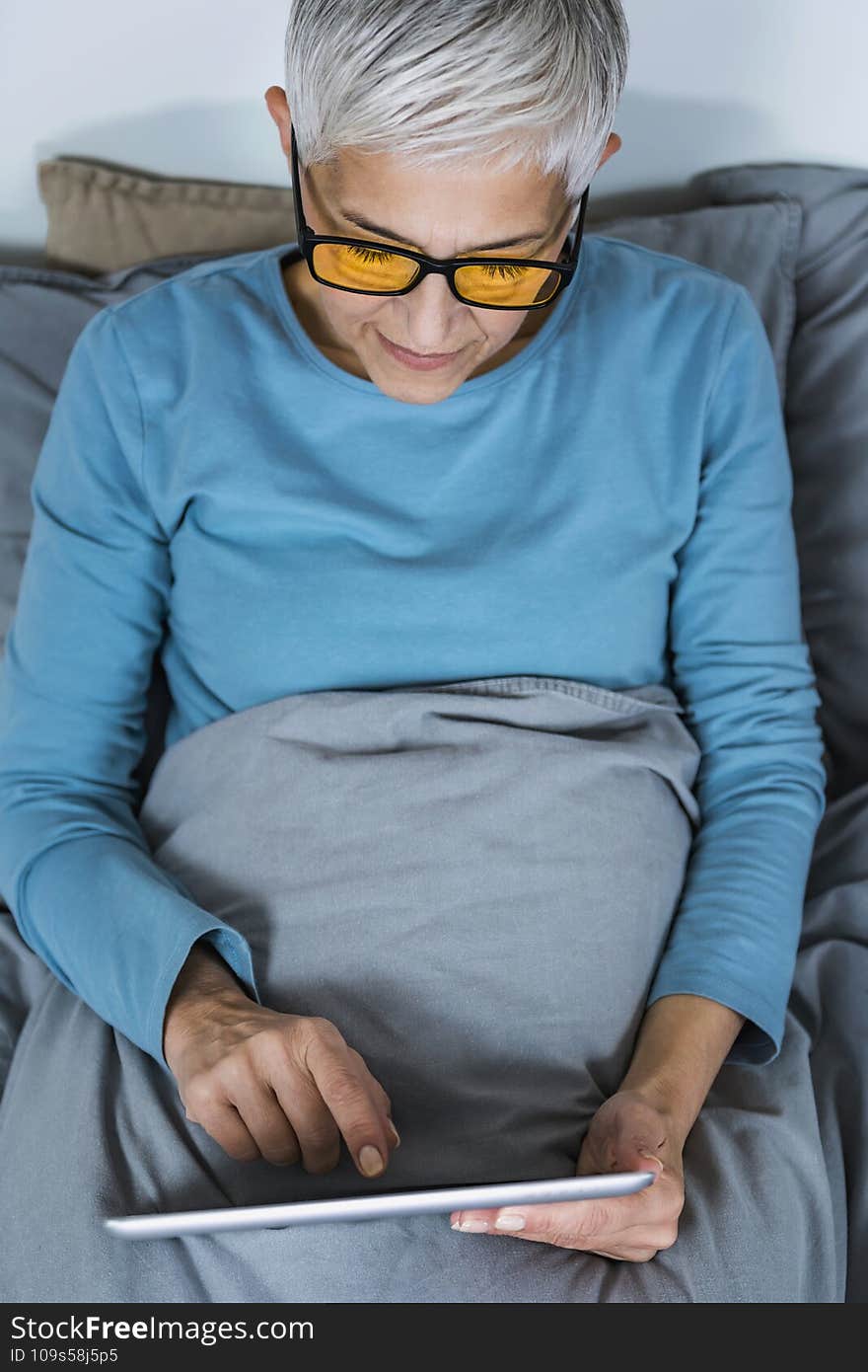 Mature woman wearing blue light blocking glasses with amber lenses, lying in bed before sleep, looking at tablet screen. Mature woman wearing blue light blocking glasses with amber lenses, lying in bed before sleep, looking at tablet screen