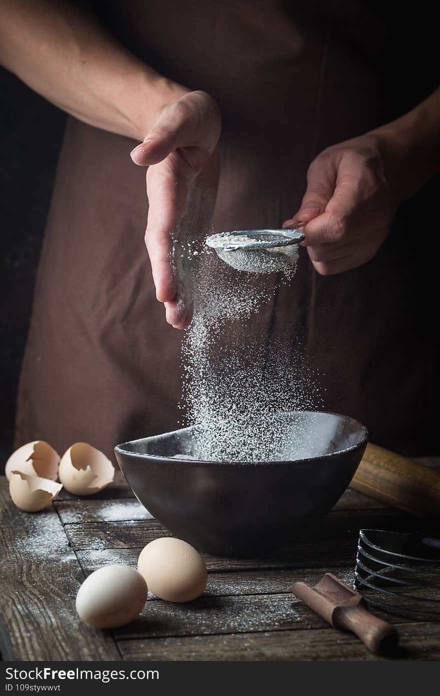 Professional chef hands sifting flour in a bowl for cooking with baking utensils over dark background. Professional chef hands sifting flour in a bowl for cooking with baking utensils over dark background