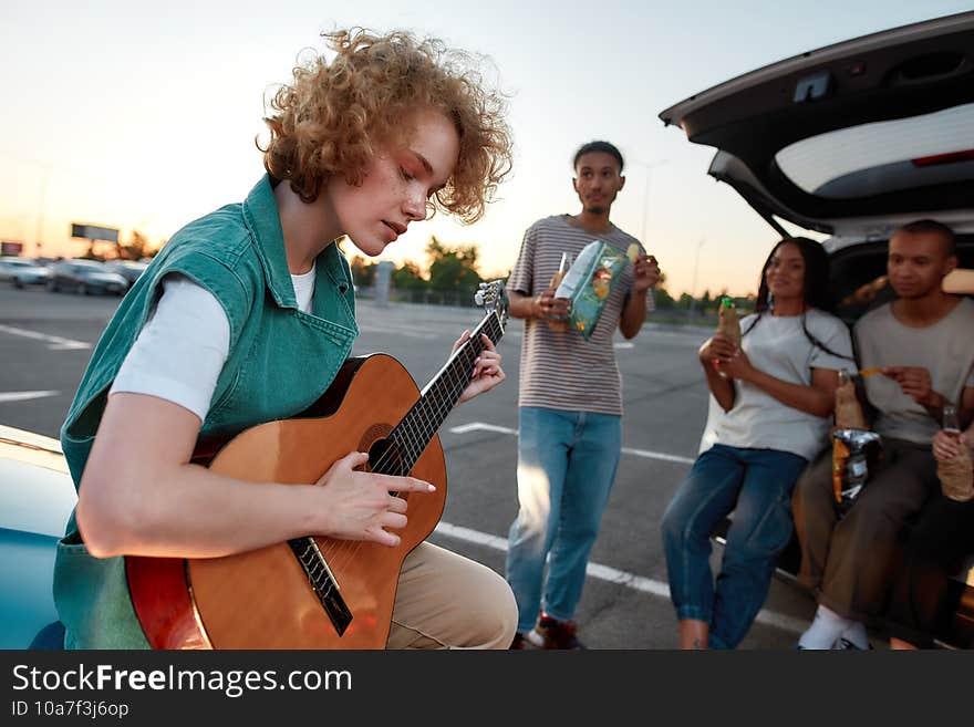 A close up of a casually dressed redhead curly young woman playing acoustic guitar outside on a parking site with her